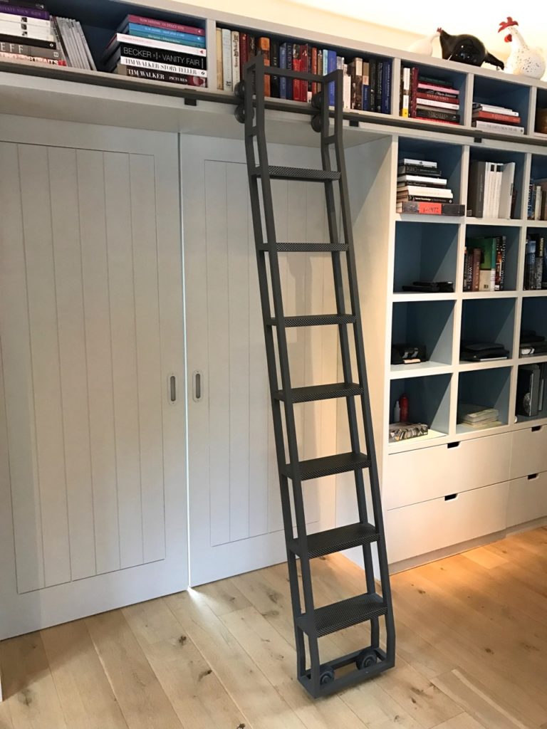 A dark gray library ladder leans against a tall bookshelf filled with books and decor items, next to closed white double doors and a set of lower drawers on a light wood floor.
