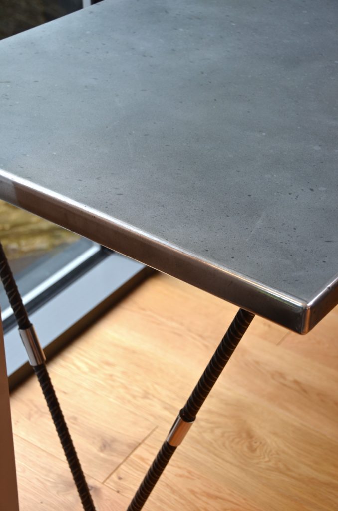 A close-up of a modern table with a smooth, zinc top and black steel legs—a perfect accent for industrial interior design—set on a light wood floor next to a window.