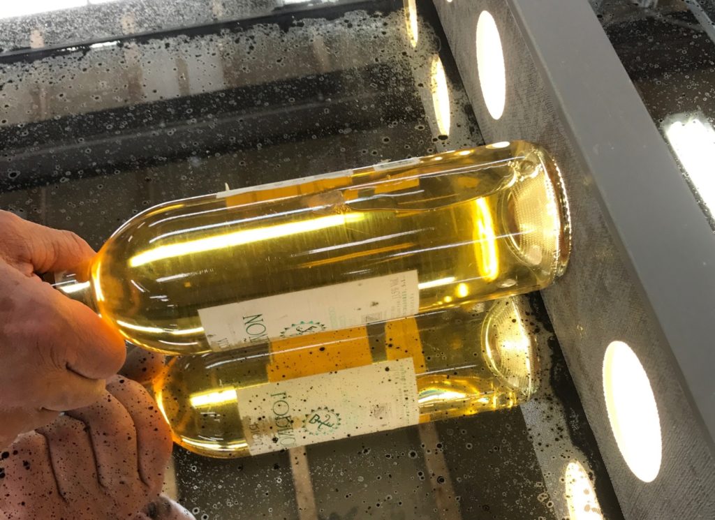 A person holds a clear glass bottle of white wine up to an illuminated bottle display for inspection, with another similar bottle reflected beneath, showing golden liquid inside.