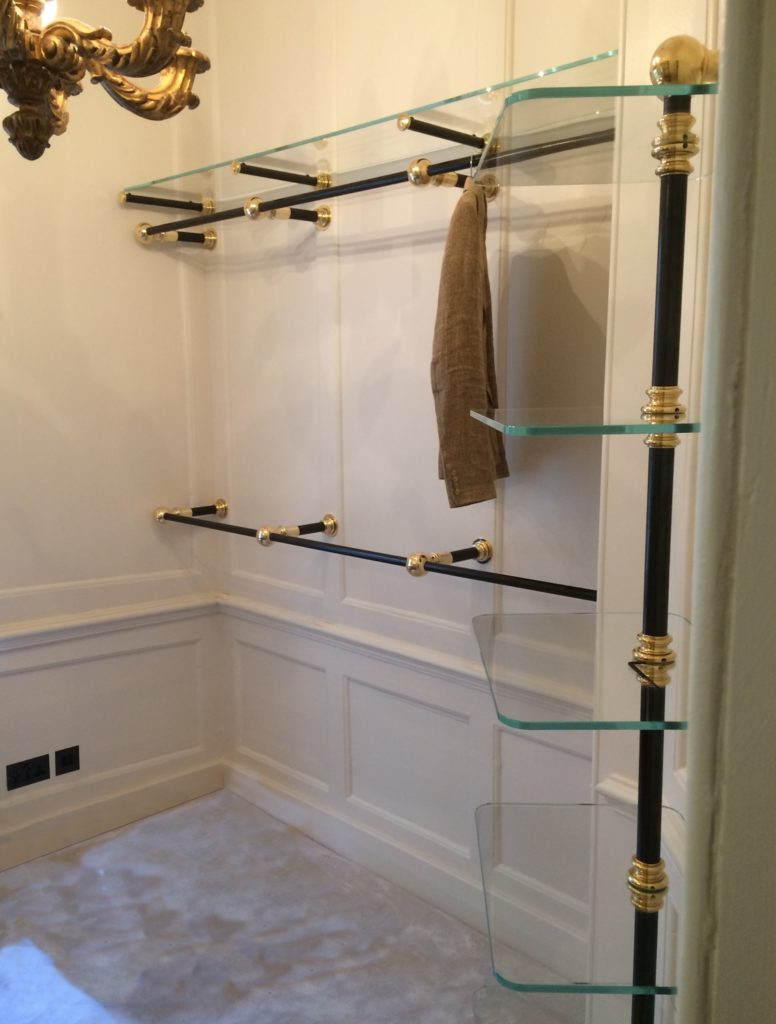 A small, elegant bespoke dressing room features glass shelves, brass and black supports, and a single brown jacket hanging. White paneled walls add sophistication, while a chandelier is partially visible in the upper left corner.