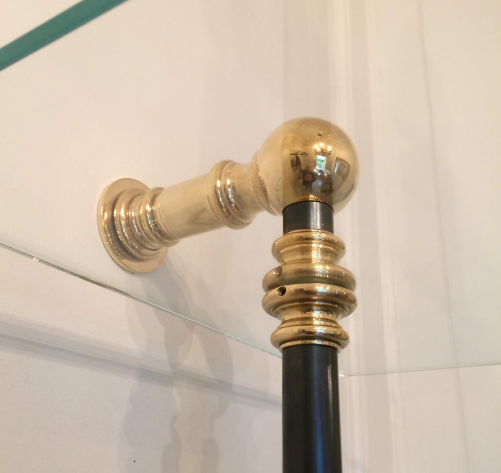 A close-up of a polished brass and black metal bracket securing a glass shelf to a white wall in a bespoke dressing room, with reflections visible on the shiny metal surface.