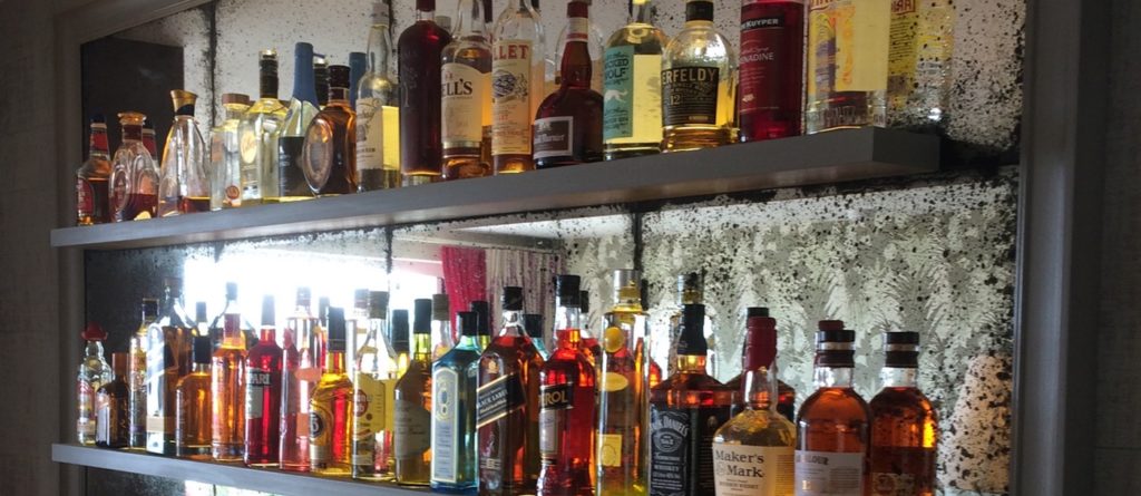 Two shelves filled with assorted bottles of liquor, including whiskey, gin, and other spirits, are showcased as an illuminated bottle display against a mirrored, slightly tarnished background. Amber and clear liquids shine invitingly within the bottles.