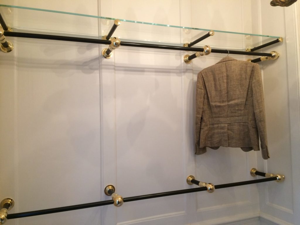 A beige blazer hangs on a black and brass clothing rod in a bespoke dressing room, mounted to a white paneled wall. The rods are empty except for the blazer, and a glass shelf is attached above.