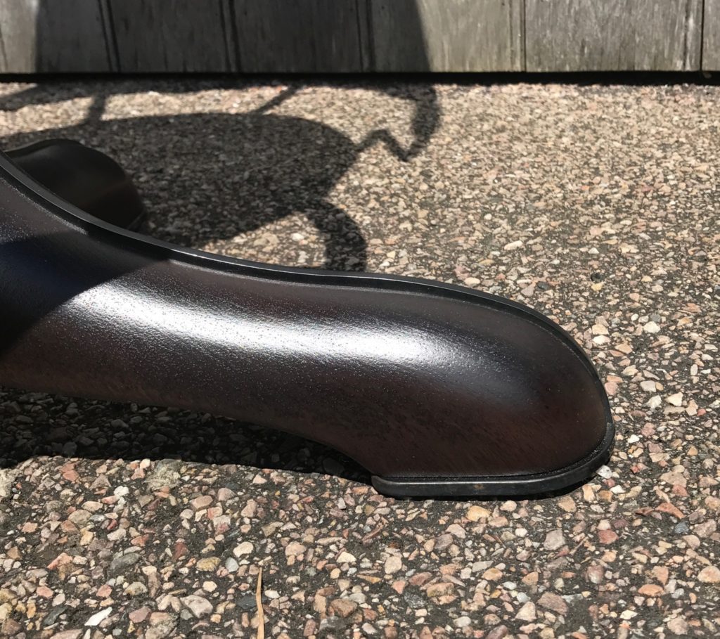 Close-up of a dark wood bar stool feet, on a speckled, pebbled concrete surface, with a wooden fence and shadow visible in the background.