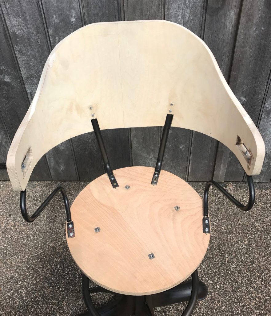 A wooden chair seat and curved backrest are partially assembled with black metal supports and screws. The chair, reminiscent of bespoke bar stools, is placed on a gravel surface in front of a wooden fence.