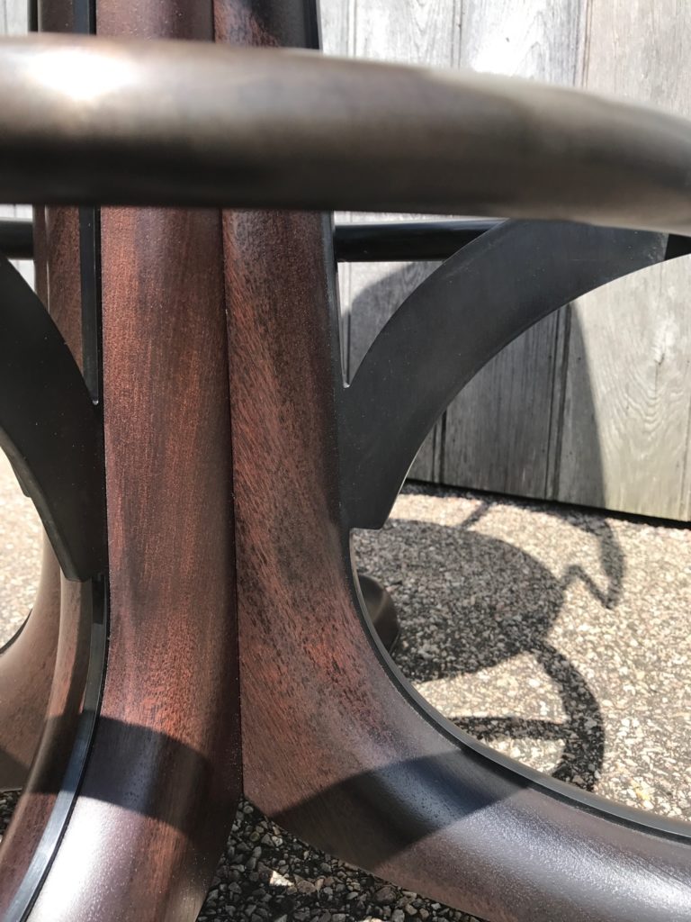 Close-up of a wooden bar stool with a dark metal ring and support brackets, set on a textured outdoor surface. Sunlight casts sharp shadows and a wooden fence is visible in the background.
