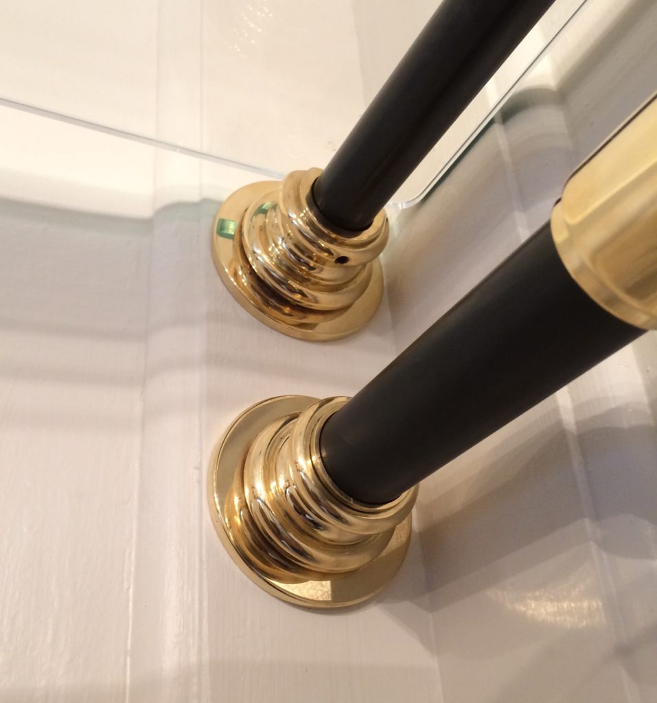 Close-up of two black vertical poles with shiny brass bases and fittings, mounted on a white surface in a bespoke dressing room near a mirror reflecting part of the scene.