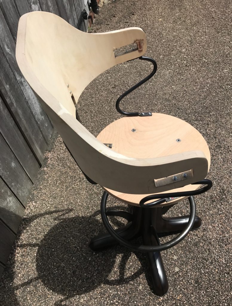 A wooden swivel chair with an unfinished seat and curved backrest, featuring exposed metal brackets and a black metal base, recalls bespoke bar stools as it sits outdoors on a rough, pebbled surface beside a wooden fence.