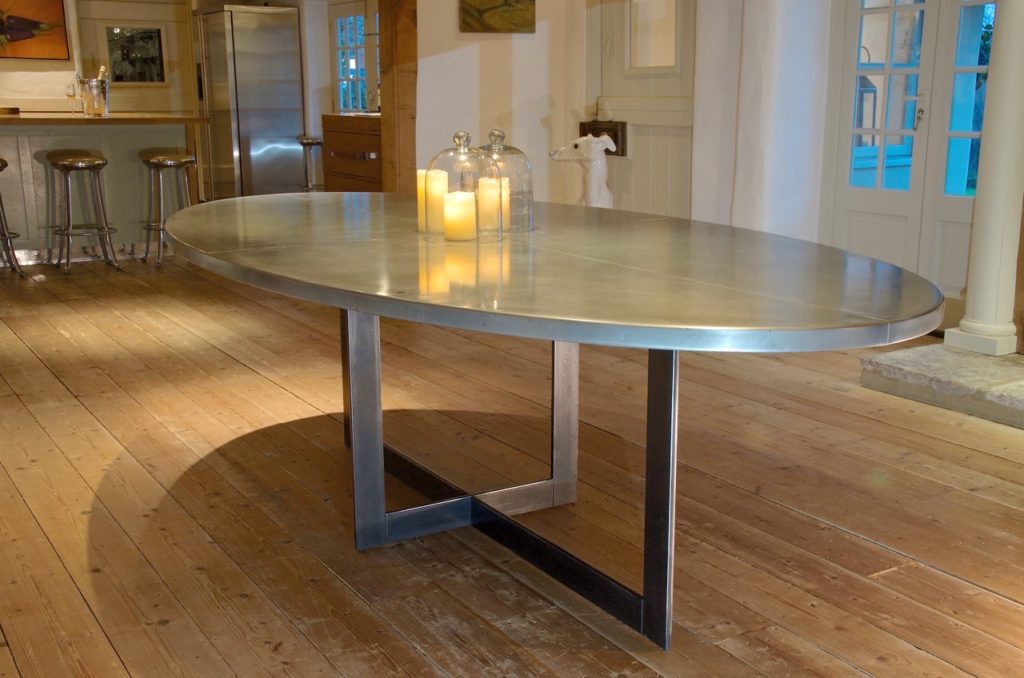 A modern oval bespoke zinc dining table with geometric legs sits on a wooden floor. Three lit candles under glass domes are placed at the centre. The background shows a kitchen area and large windows with natural light.