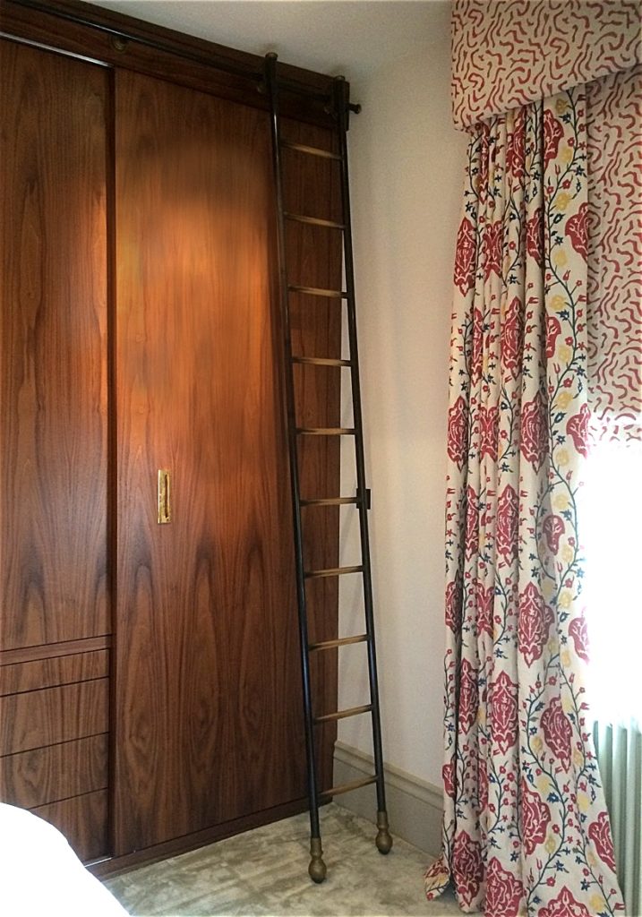 A tall wooden wardrobe with sliding doors stands beside a window with colourful red and blue patterned curtains; a dressing room ladder leans against the wardrobe.