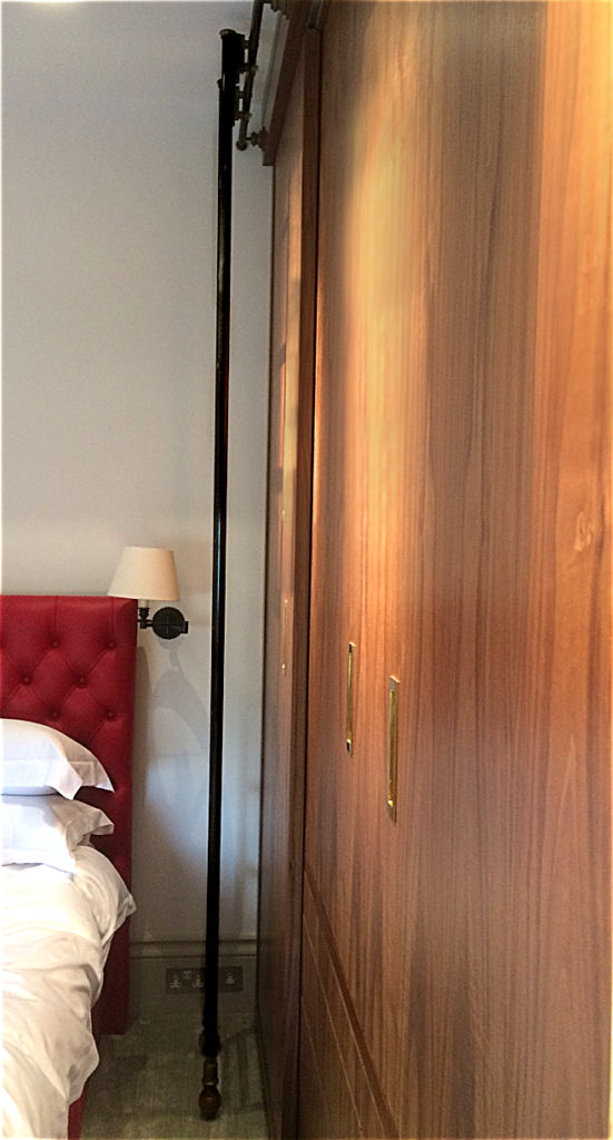A tall dressing room ladder stands between a red upholstered bed and a large wooden sliding wardrobe in a bedroom with white walls and a wall-mounted lamp.