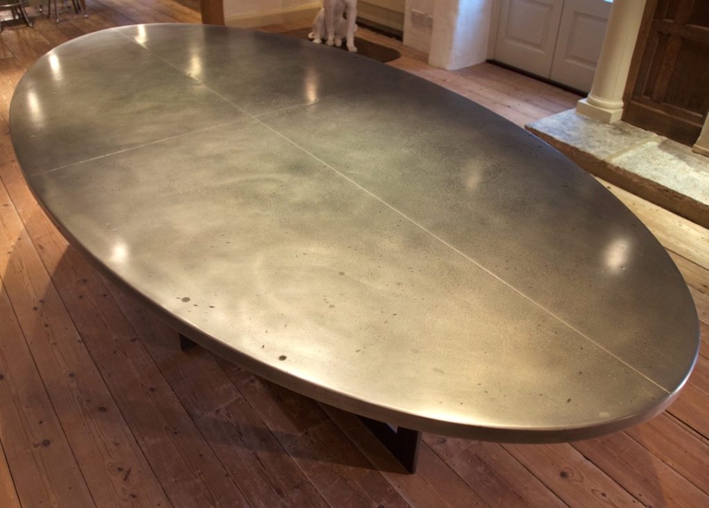 A bespoke oval dining table with a smooth, metallic, and reflective surface sits gracefully on a wooden floor in a bright room.