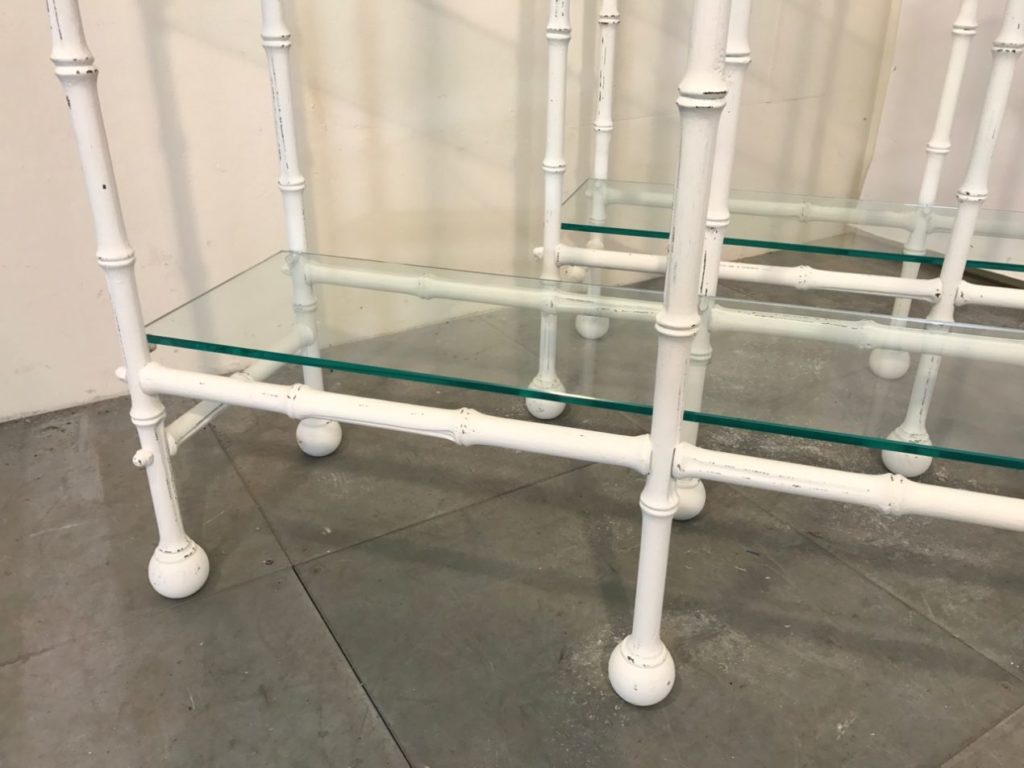 A close-up view of a shelving unit with white metal legs resembling bamboo, round feet, and a clear glass shelf. Reminiscent of classic solid brass étagères, the unit is set on a grey concrete floor against a light-coloured wall.