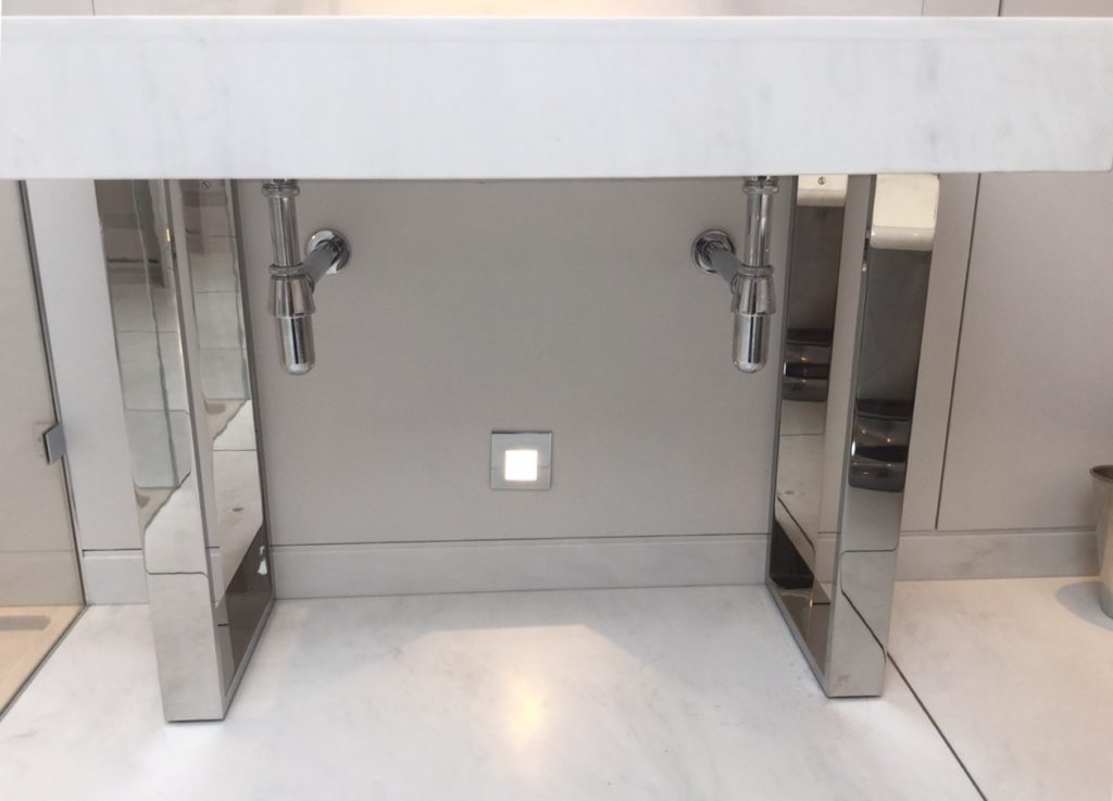 A modern bespoke bathroom washstand features exposed chrome pipes and mirrored zinc side panels, set against a light wall and floor. A square electrical socket is conveniently placed beneath the sink.