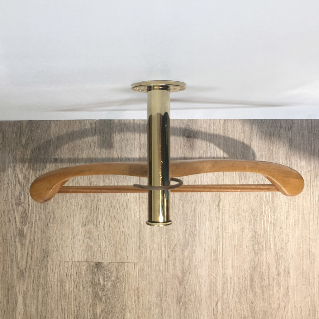 A wooden coat hanger with a brass hook is hanging from the Retractable Wall Mounted Valet Hook, which is attached to a brass fitting on a light wood wall and casts a shadow beneath it.