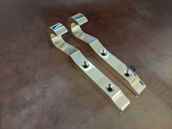 A pair of Solid Brass Ladder Hooks for hook-on ladders with a curved shape and mounting holes are shown side by side on a brown, textured surface.