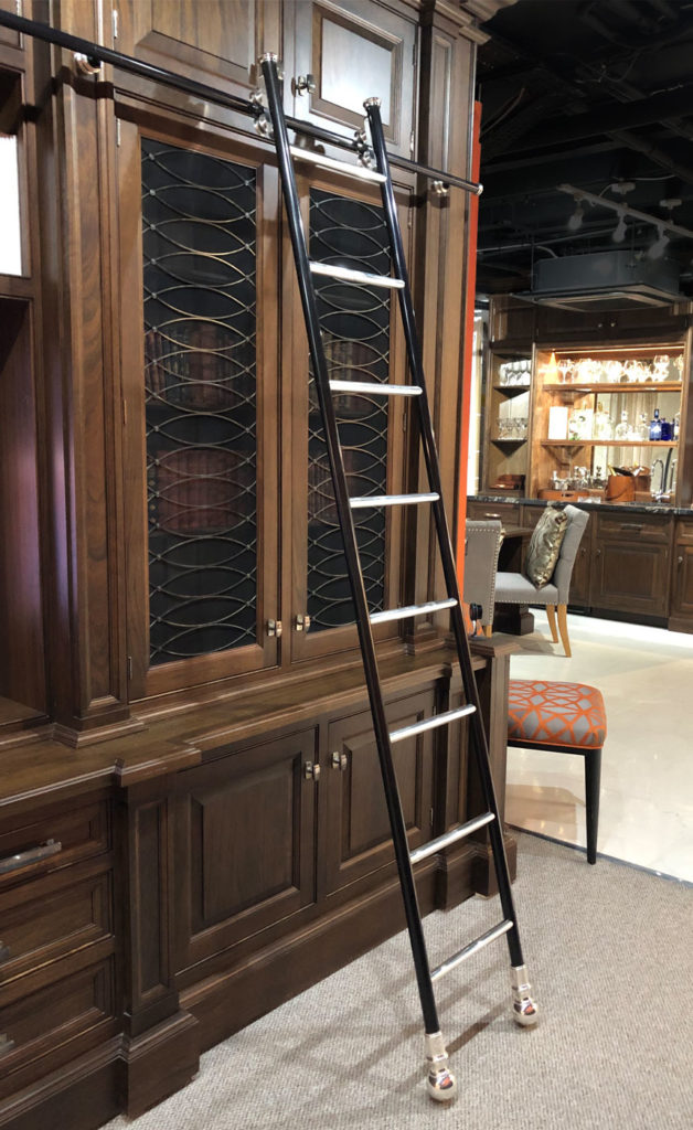 A rolling library ladder is leaning against a tall, dark wooden bookcase with glass cupboard doors. In the background, a dining area with chairs and a table is visible.