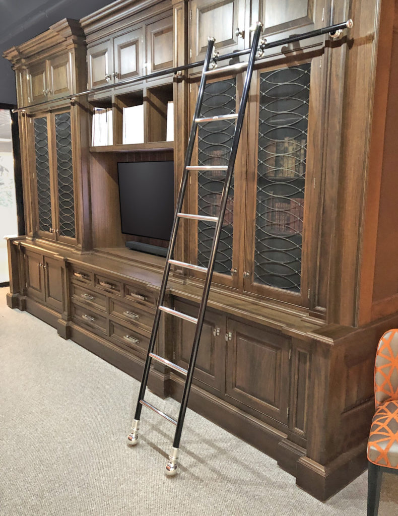 A tall, dark wood built-in bookcase with glass cupboard doors, drawers, and a mounted television. A rolling ladder is attached to a rail, providing access to higher shelves. An orange patterned chair is partially visible on the right.