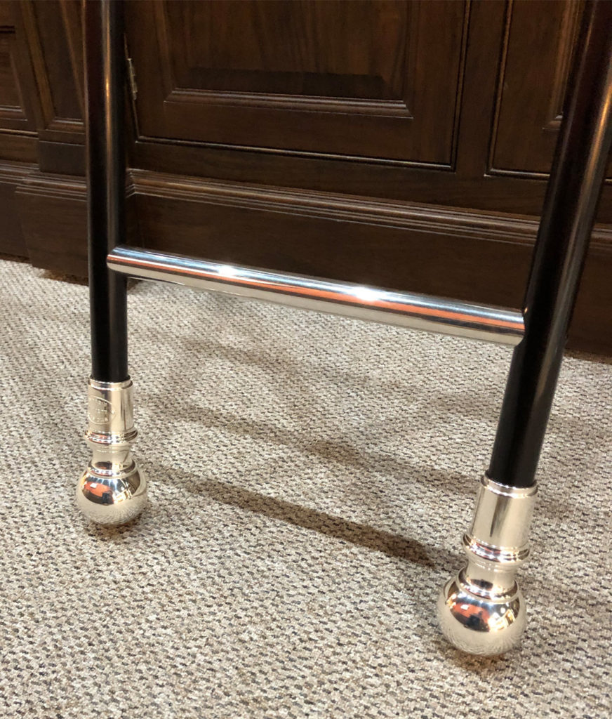 Two black legs of a rolling library ladder, each with a shiny silver ball-shaped foot, are connected by a horizontal silver rung. The ladder stands on a light-coloured carpet in front of a dark wooden cupboard.