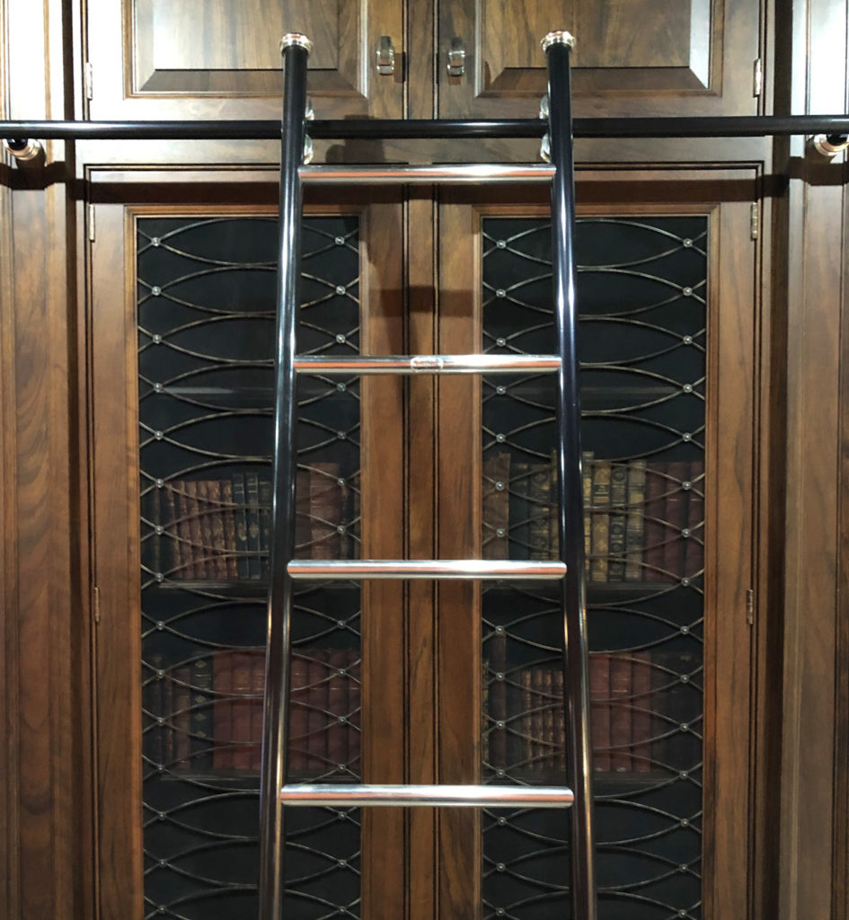 A rolling library ladder leans against a tall, dark wooden bookcase with glass doors, behind which old books with patterned spines are visible. The top of the ladder reaches cupboard doors above the shelves.