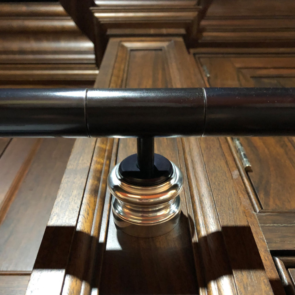 A black metal rod for a rolling library ladder is mounted on a shiny metallic bracket attached to a wooden panelled wall. Warm lighting highlights the wood grain and metallic surface.