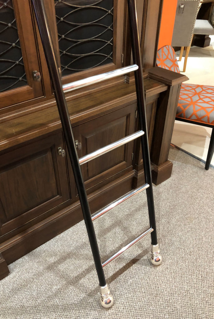 A close-up of a dark metal rolling library ladder with silver rungs, leaning against a wooden cupboard. Part of an upholstered chair with an orange geometric pattern is visible in the background.