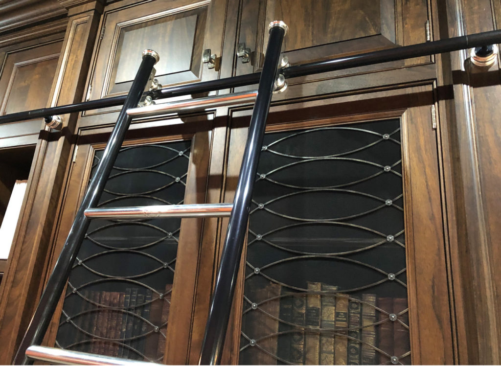 A black rolling library ladder with silver steps is attached to a track in front of tall, wooden bookcases filled with books and glass-fronted cabinet doors.