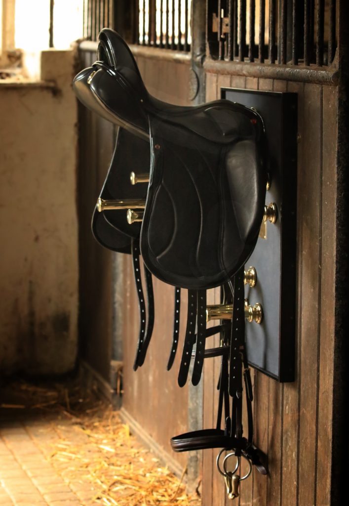 A black leather horse saddle hangs neatly on a wall-mounted tack rack in a stable, with sunlight filtering through the stable door and straw scattered on the brick floor below.