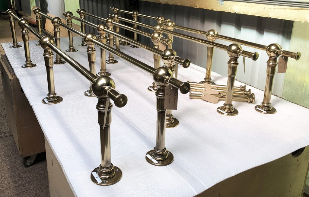 Several polished brass railings with mounting bases are lined up on a padded white surface, reflecting light in an industrial or workshop setting—ideal components for bespoke dressing room furniture. A pair of gloves is placed in the background.