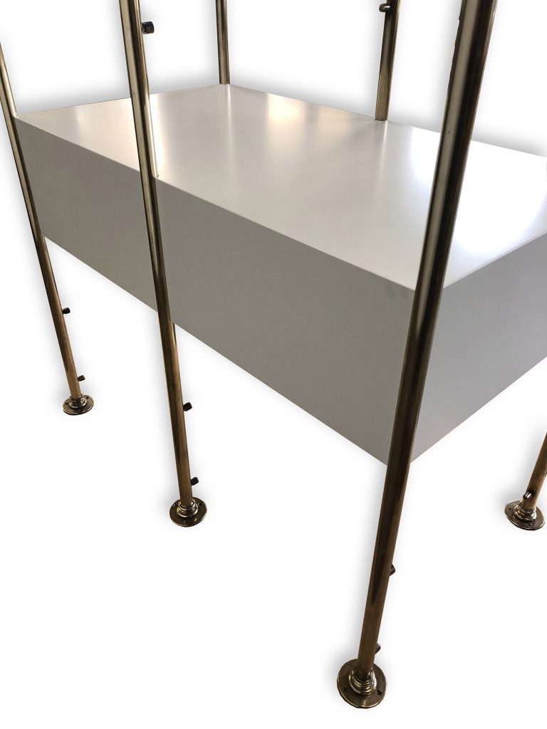 A white rectangular dressing room furniture, is supported by four shiny brass poles with round bases, set against a plain white background.