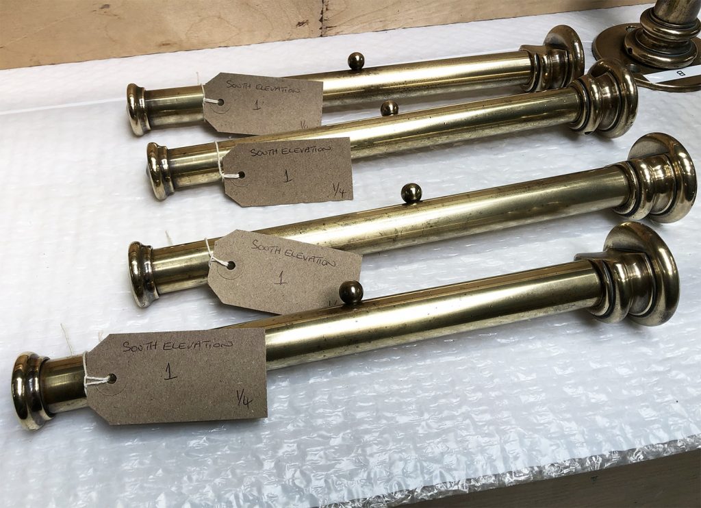 Four brass rods with finials, each tagged with a brown label reading South Elevation 1 1/4, are arranged on a bubble wrap surface, evoking the precision and elegance often found in bespoke dressing room furniture.