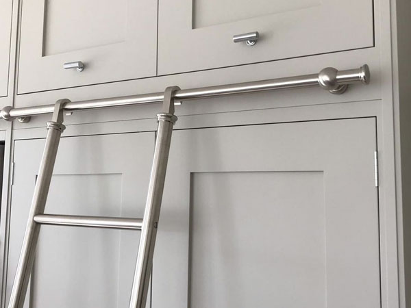 A brushed nickel hook-on library ladder complements the light gray cabinets, with a sleek, modern design. Attached to horizontal rails, it allows easy access to higher cabinet sections. The ladder's silver finish enhances the elegance of the dual-door cabinetry and drawer handles.