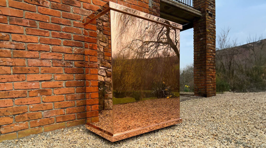 A polished brass drinks cabinet stands outdoors, reflecting a tree and the surrounding landscape. It rests on gravel next to a brick wall, with a partially visible building and overcast sky in the background.