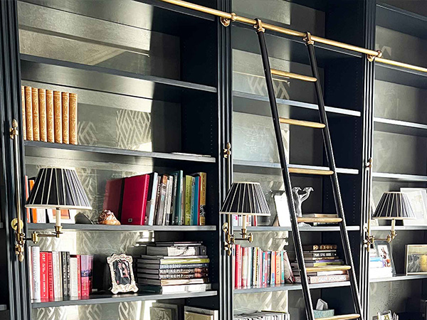 A bespoke black bookcase, crafted from patinated brass, houses an array of books and decorative items. It features two wall-mounted lamps with striped shades and a rolling library ladder attached to sleek rails for easy access to the upper shelves.