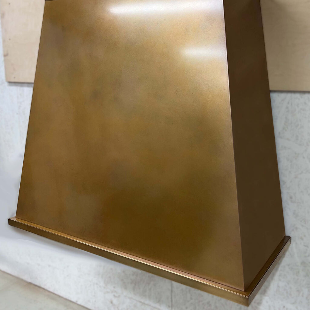 Mounted against a light-colored wall, the Handmade Patinated Copper Cooker Hood is a large piece with a smooth and reflective pyramid shape that adds an elegant touch to the kitchen space.
