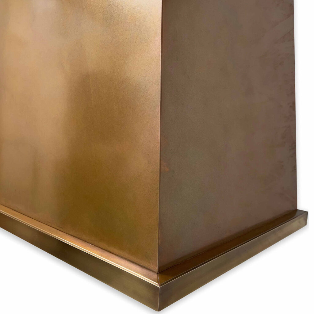 A close-up view of the Handmade Patinated Copper Cooker Hood showcases its smooth, reflective surface with a warm, golden hue. This triangular-shaped hood, featuring a flat, broad base, highlights its metallic allure through masterful craftsmanship using patinated copper.