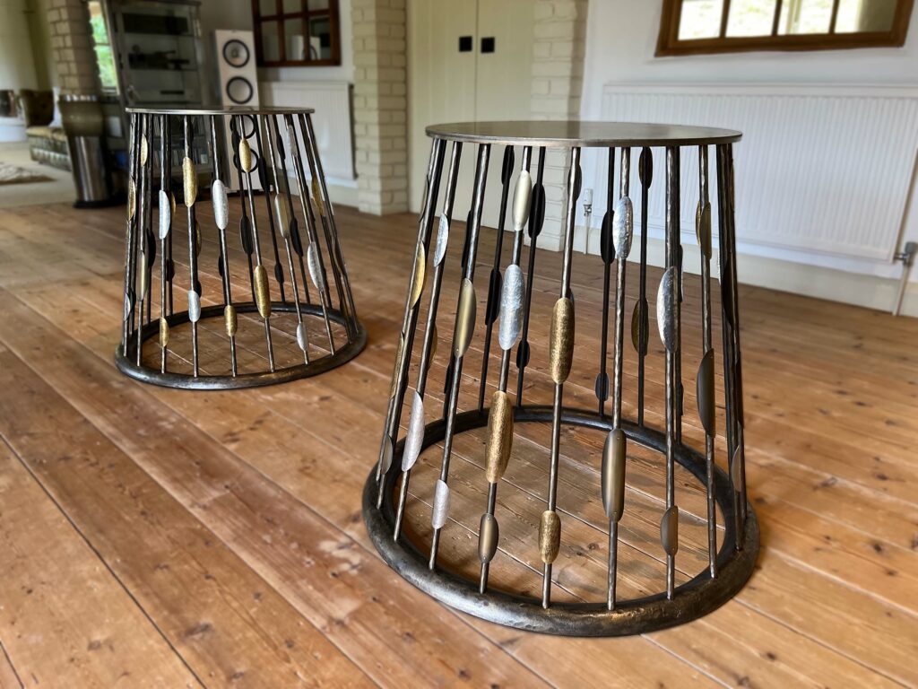 Two artistic steel table bases with wire frames and leaf-shaped accents in gold and silver hues stand on a wooden floor. A minimalist interior setting with cream brick walls and large windows complements the design.