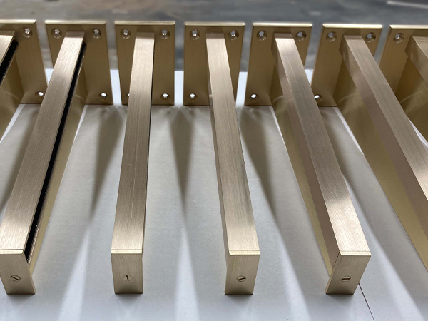 Close-up of several brass-colored rectangular metal bars with holes, aligned in a row on a flat surface. Each bar is attached to a flat bracket at one end, suggesting they might be components for back bar shelving or other fixtures.