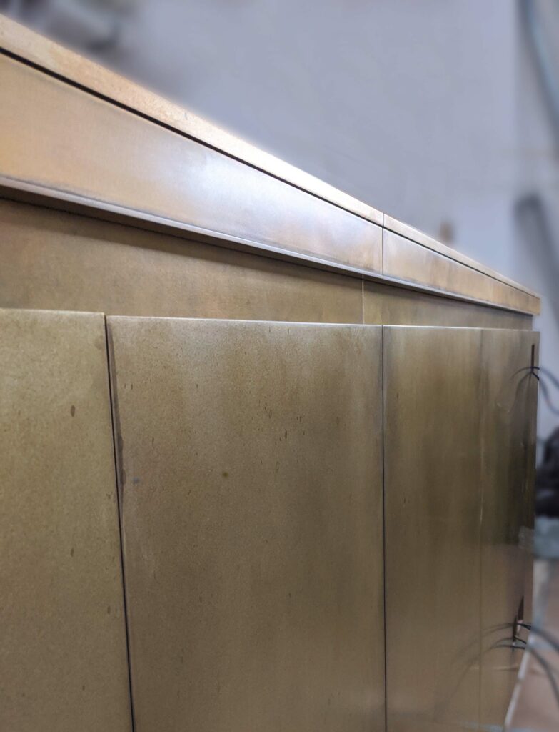 Close-up view of a metallic cabinet with a slightly reflective surface, complemented by wall panelling that adds depth to its design. The cabinet features a series of horizontal panels and subtle vertical lines, enhancing its modern and sleek appearance. The background is softly blurred.