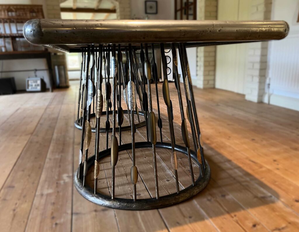 A unique zinc table with rounded edges and custom steel table bases composed of vertical metal rods resembling arrows. The table sits on a wooden floor in a room with light-colored brick walls.
