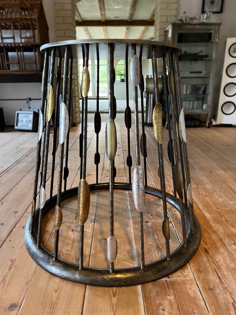 A metal circular coffee table features vertical bars embellished with metallic oval shapes in gold, silver, and bronze hues. Its custom steel table base sits elegantly on a wooden floor, complementing the modern interior in the background.