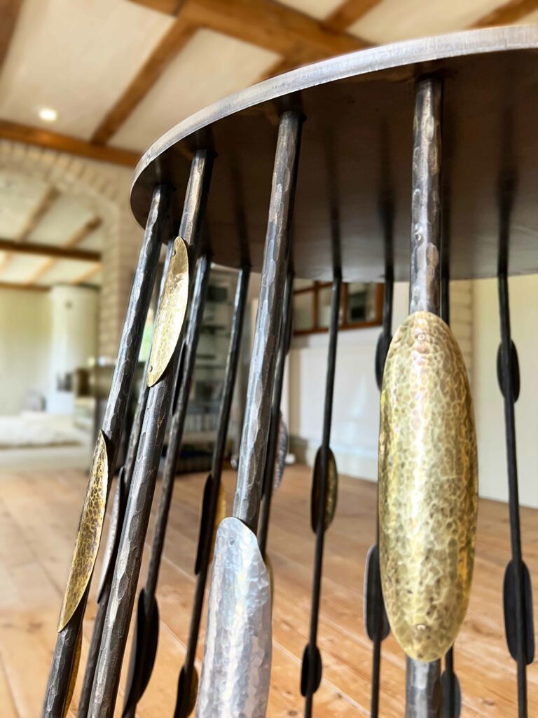 Close-up of a steel and brass table with vertical rods and decorative gold and silver oval elements, supported by custom steel table bases. The background shows a room with wooden beams, light walls, and a distant couch, creating a warm, rustic atmosphere.