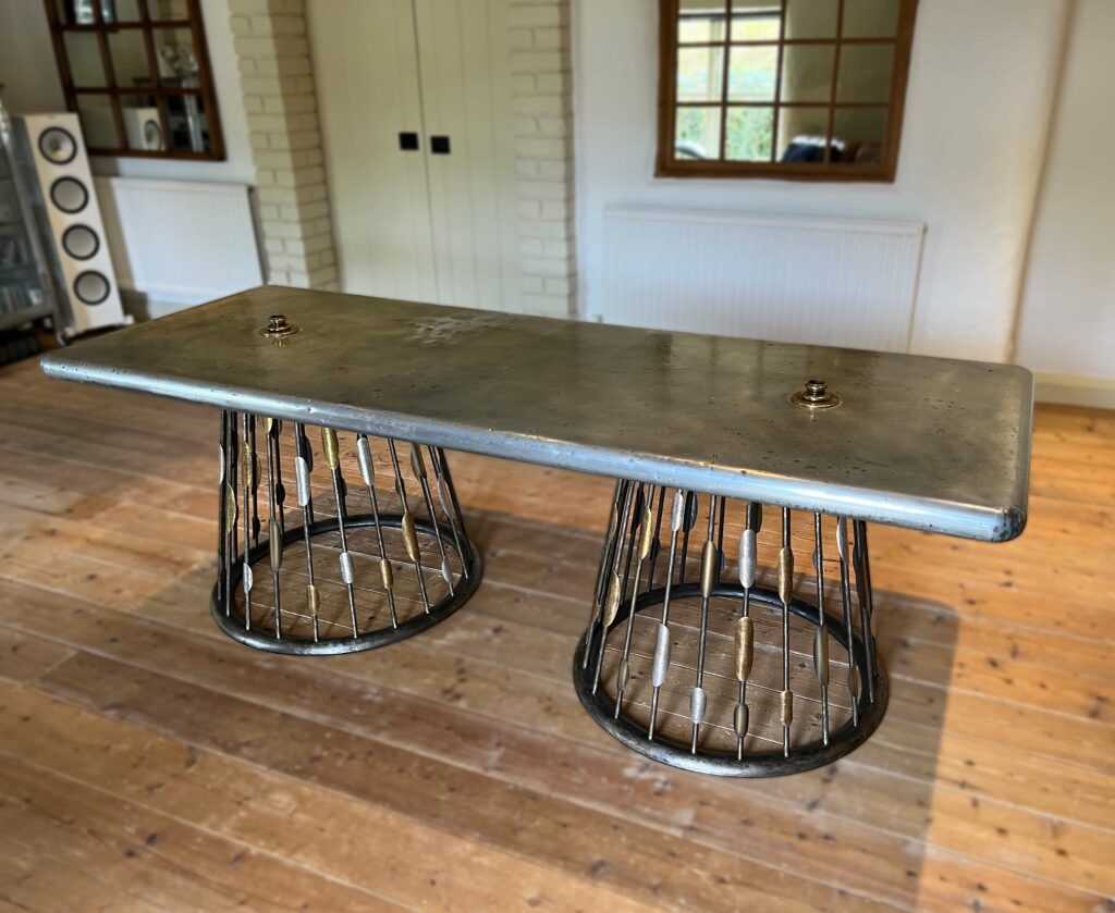 A large, industrial-style table with a metallic top and custom steel wireframe bases stands on a polished wooden floor in a room with white brick walls and windows.
