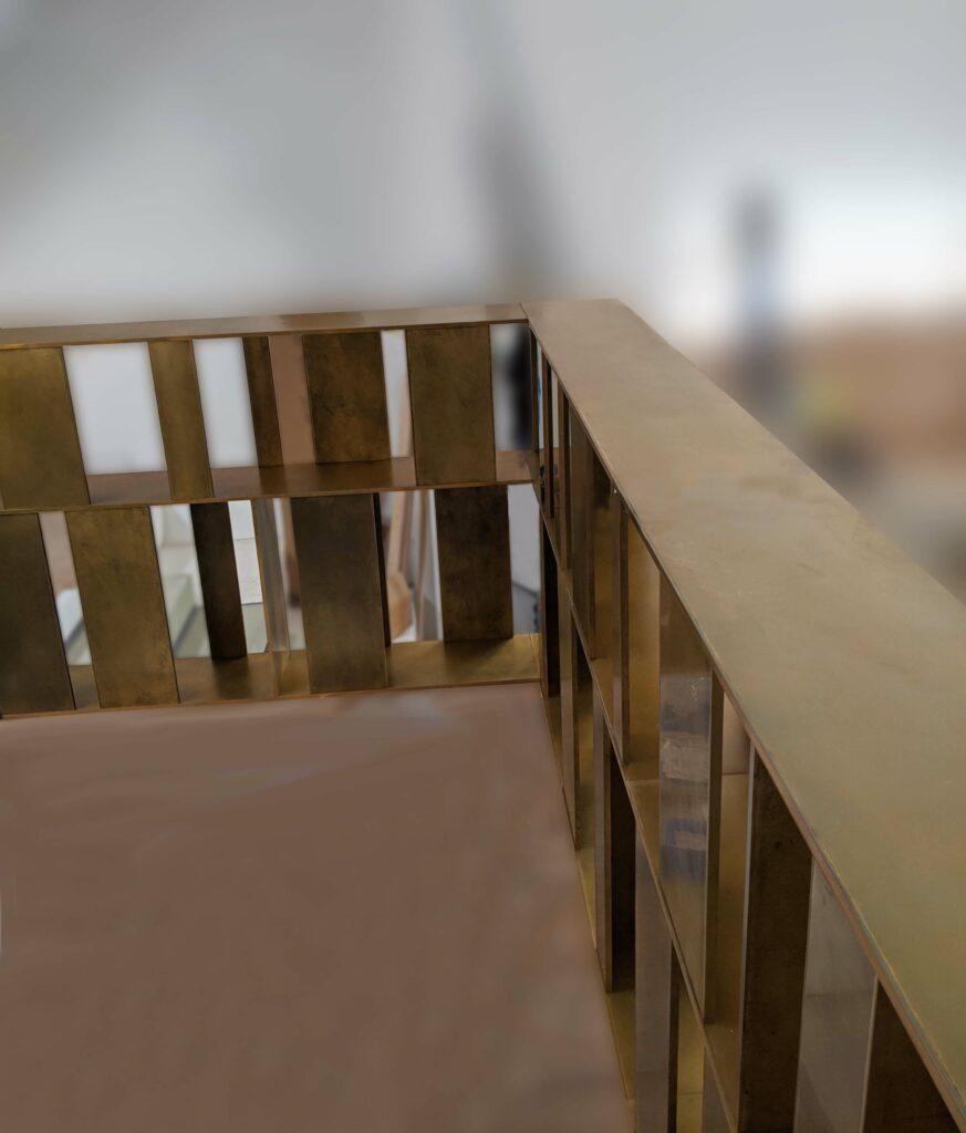 A sleek, modern metal railing with vertical slats and a smooth top rail stands like an elegant room divider in a softly lit environment. The background is blurred, creating a minimalist and contemporary aesthetic.