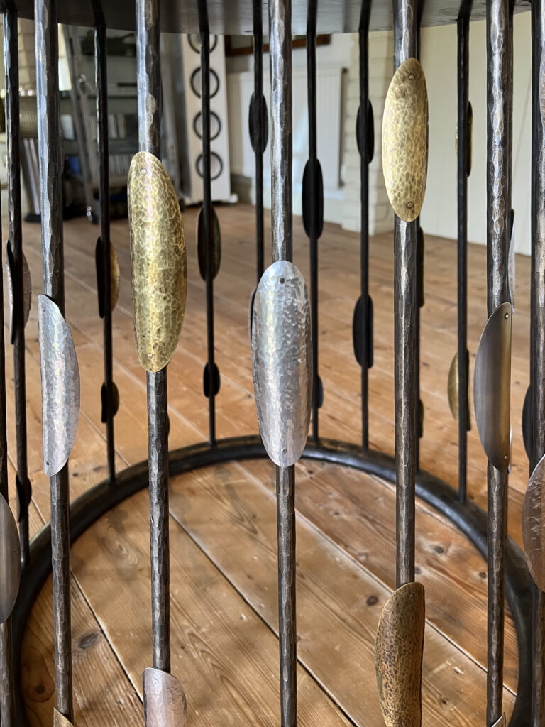 Close-up of a custom steel table base featuring artistic vertical rods and decorative, leaf-shaped metal accents in silver and gold finishes. Set on a wooden floor, with a blurred room interior providing the backdrop.