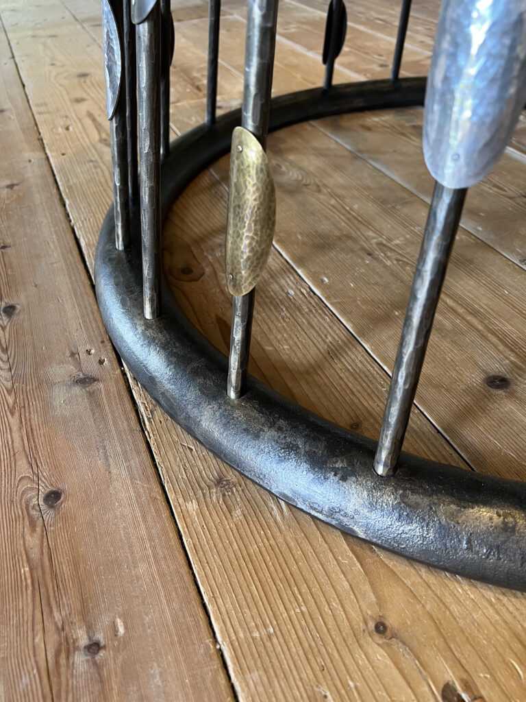 Close-up of a custom steel table base on a wooden floor, with vertical rods attached intermittently, some featuring textured, oval-shaped metallic decorations.