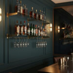A stylish home bar with dark teal walls features brass back bar shelving, two brass-framed glass shelves displaying liquor bottles, and hanging glassware beneath. Warm wall sconces illuminate the modern, elegant space.