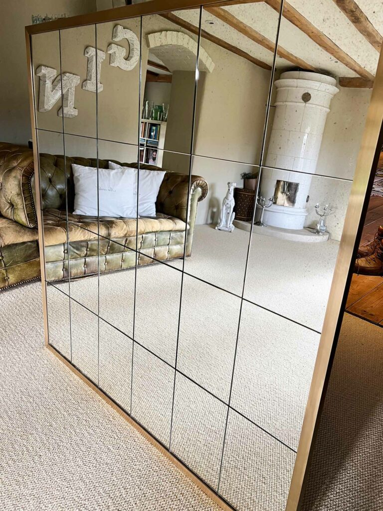 An antique mirror with a brass frame and grid pattern reflects a cozy living room, featuring a vintage leather sofa, a white ceramic stove, and rustic wooden beams on the ceiling. The flooring is carpeted in a neutral color.