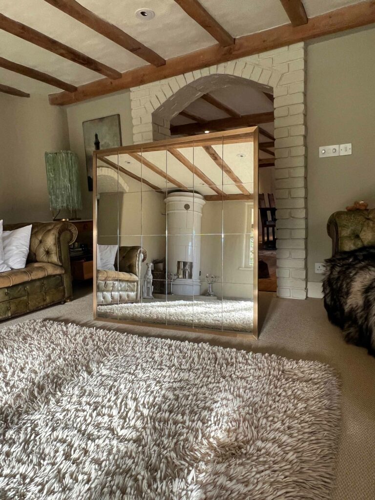 A cozy living room features an antique mirror with a brushed brass frame reflecting a white stove and wooden beams. Plush green armchairs, a shaggy beige rug, and a textured wall complete the setting. Soft lighting adds warmth to the rustic decor.