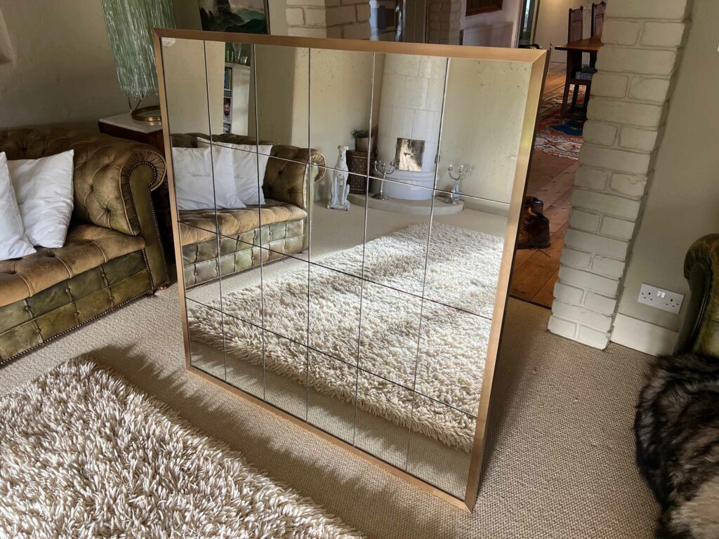 A large living room mirror with a grid design reflects a cozy space. The room features a green tufted sofa, white pillows, a plush beige rug, and a brick fireplace in the background.